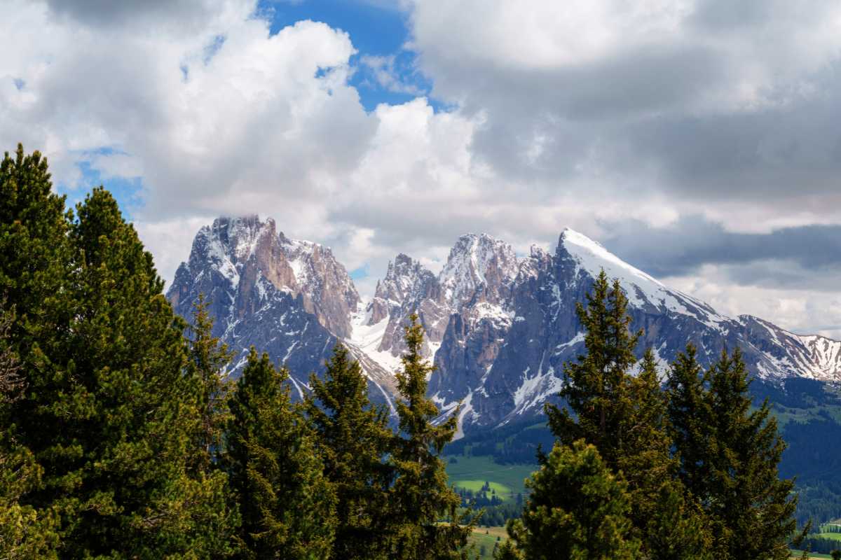 Veduta panoramica estiva di Ortisei: il paese incastonato tra i prati verdi e le Dolomiti illuminate dal sole, persone che camminano lungo il centro storico fiorito