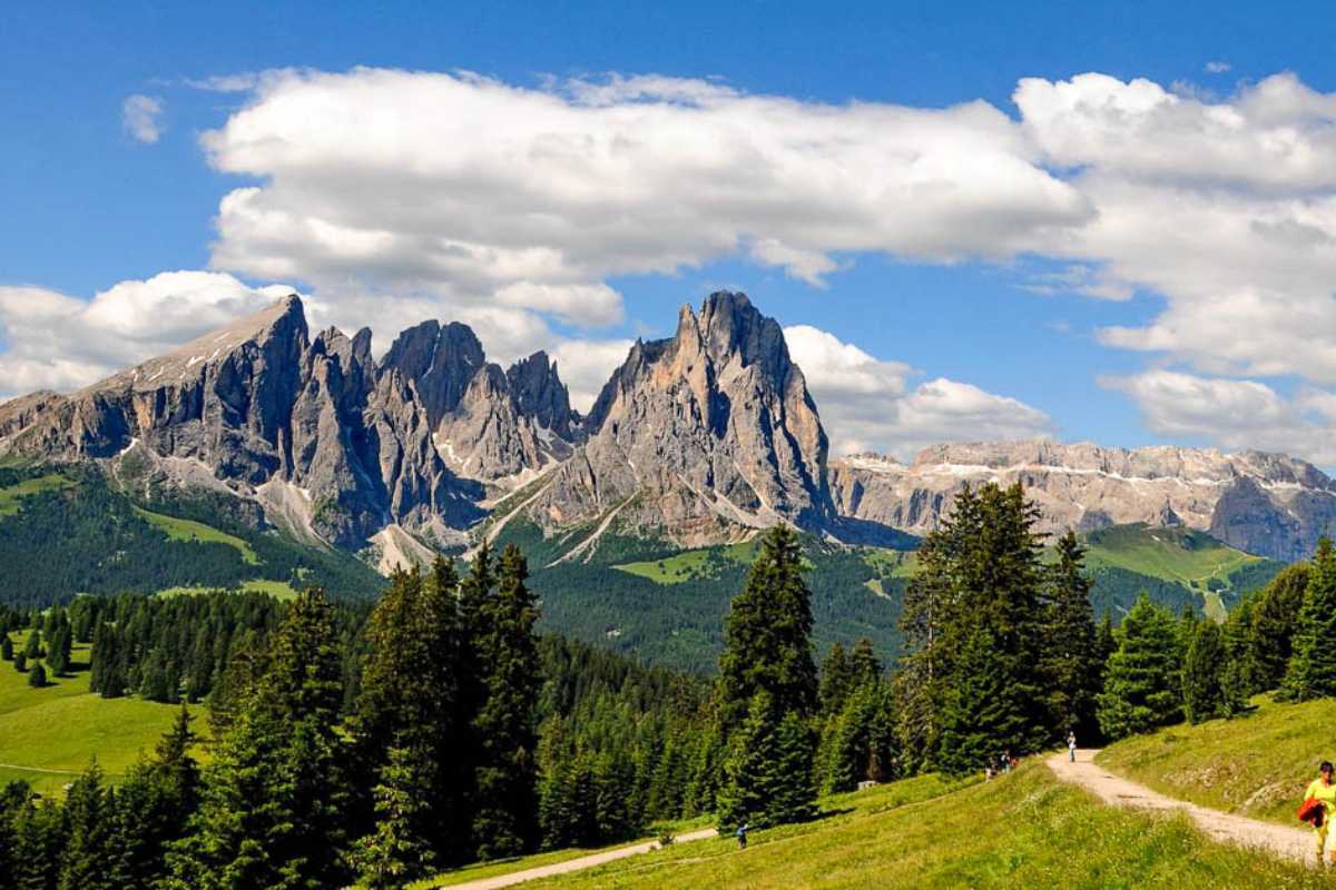 Escursionisti in estate lungo un sentiero panoramico sopra Ortisei, prati verdi, cielo azzurro, vista sulle Dolomiti e sul paese